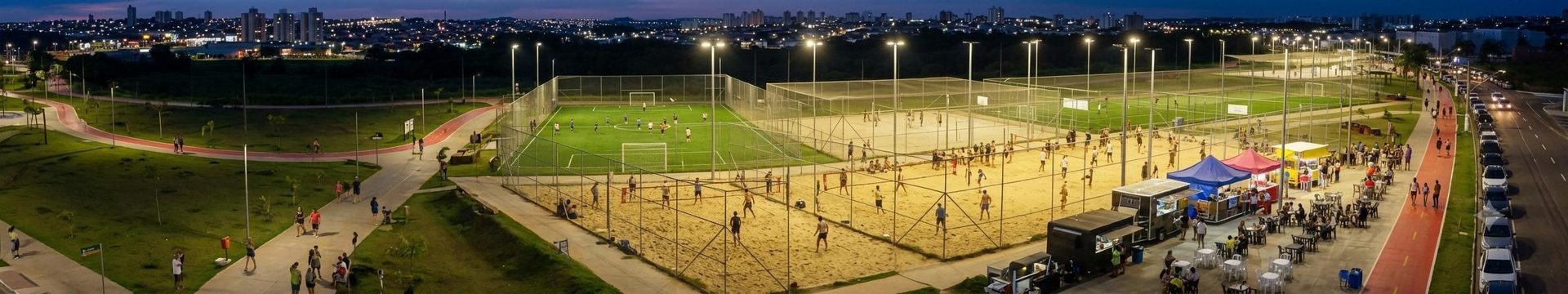 Vista aérea noturna do Lago da Fé em Hortolândia, mostrando as quadras de esportes iluminadas, pista de caminhada e food trucks com a cidade ao fundo.