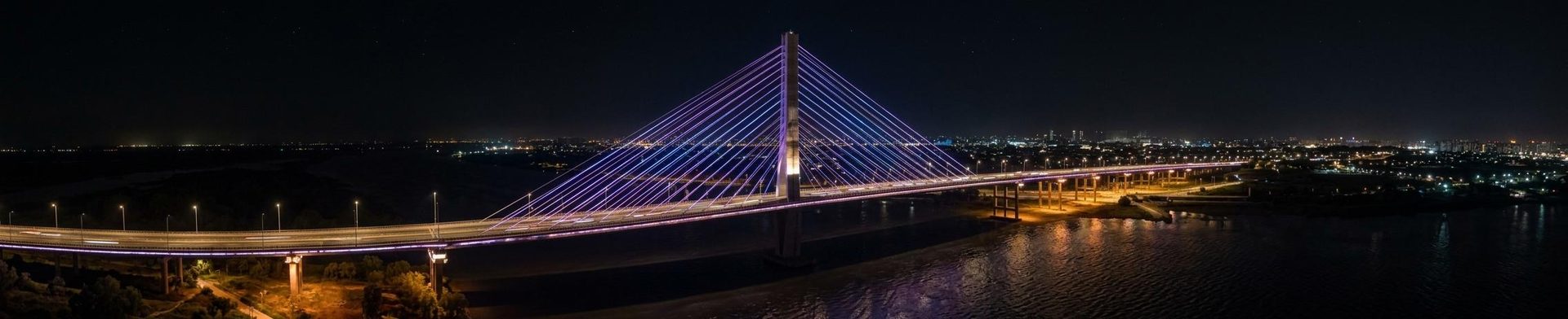 Fotografia panorâmica noturna da Ponte Estaiada de Hortolândia com suas luzes refletidas no rio e um céu estrelado.